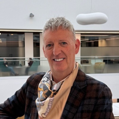 Smiling gray-haired man in a plaid blazer and patterned scarf poses in a bright, modern airport or atrium with sleek architectural details.