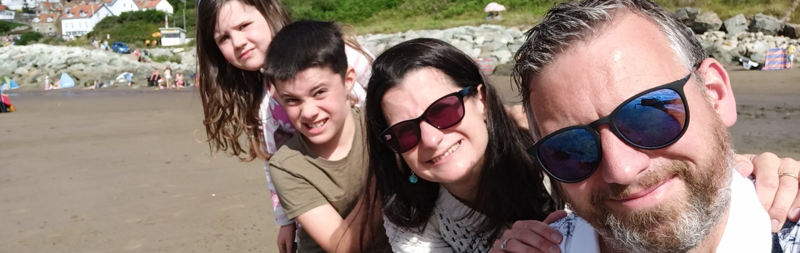 Group of people posing on a sandy beach with rocky hills and houses in the background under a clear blue sky.