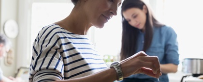 Woman in striped shirt cooking at a bright kitchen counter, focused and smiling slightly. Another woman in the background is seated, smiling contently.