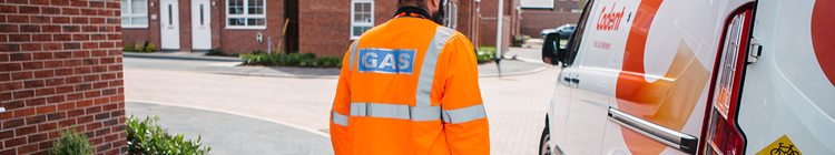 Gas worker in bright orange uniform with safety stripes seen from the back, standing near a gas company van in a residential area with brick houses.