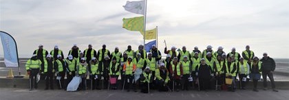 A group of people wearing high-visibility vests, holding litter pickers, pose outdoors under cloudy skies. Several flags are raised behind them.