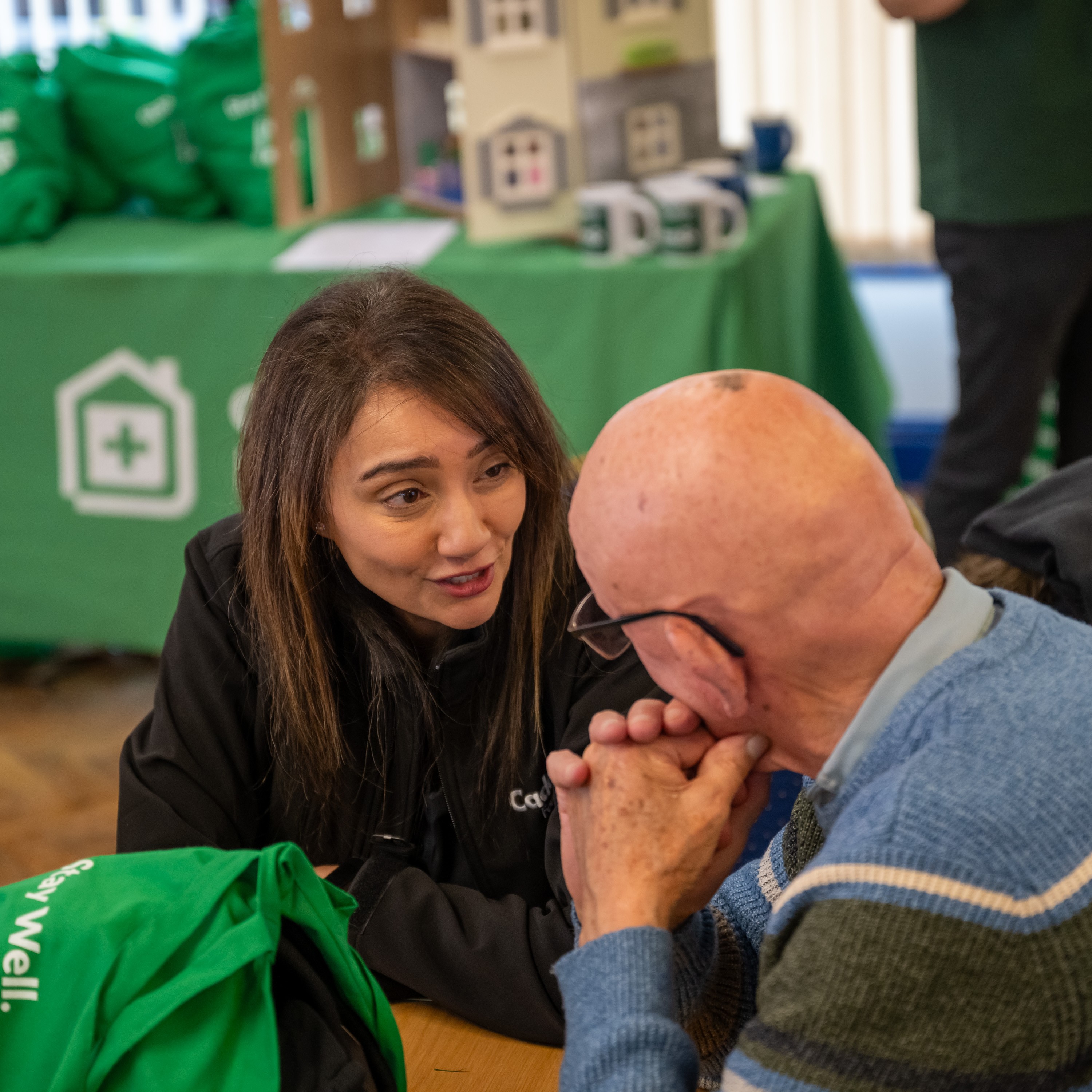 A woman and elderly man converse warmly at a community event. The table holds green bags and brochures, creating an inviting atmosphere of connection.