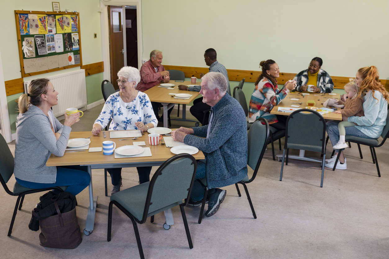 A warm gathering in a community hall features diverse people of various ages joyfully chatting and drinking coffee around tables, creating a lively and friendly atmosphere.