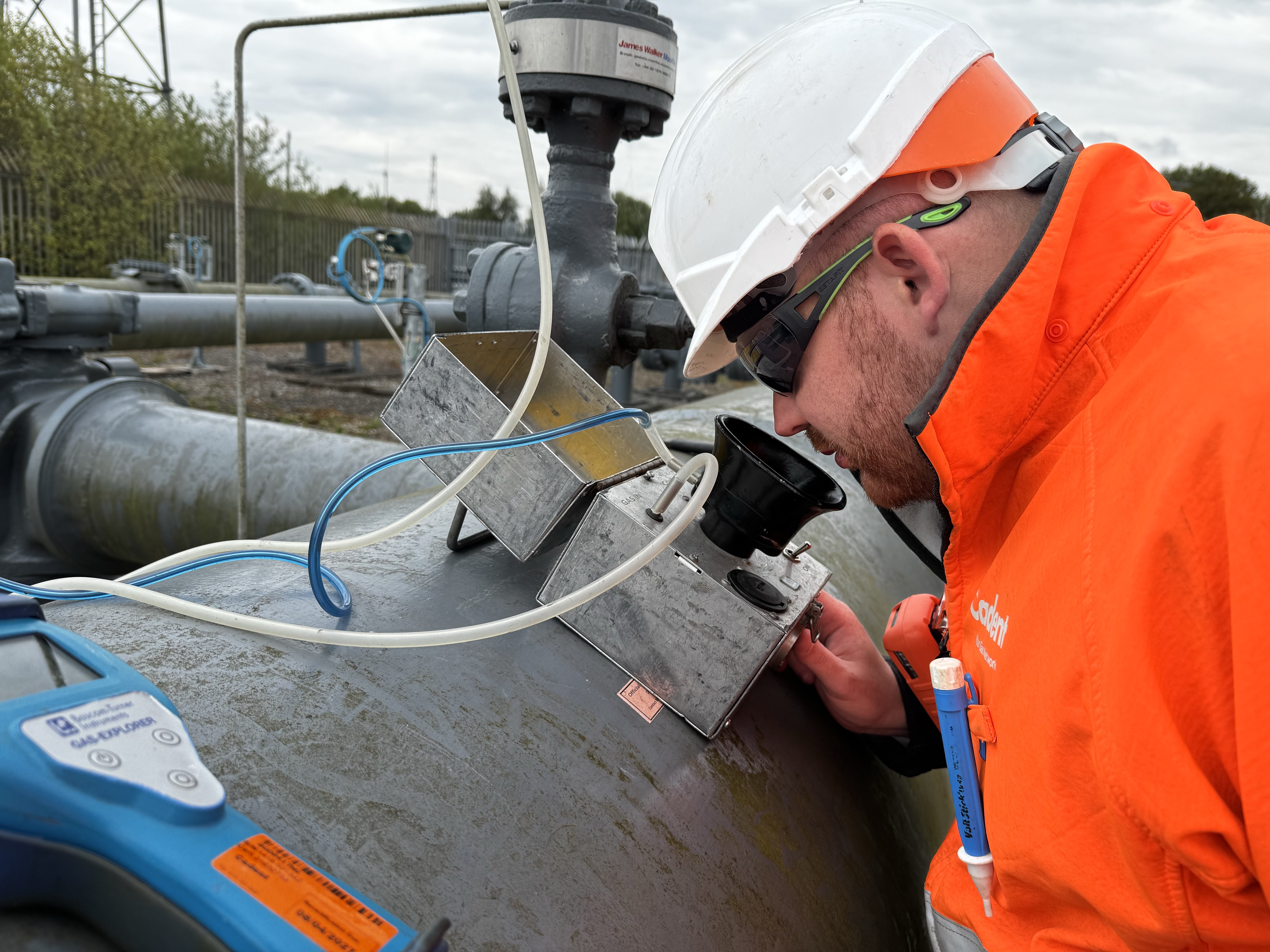 Worker in orange safety gear and hard hat inspects a large industrial pipeline with a handheld device at an outdoor facility.