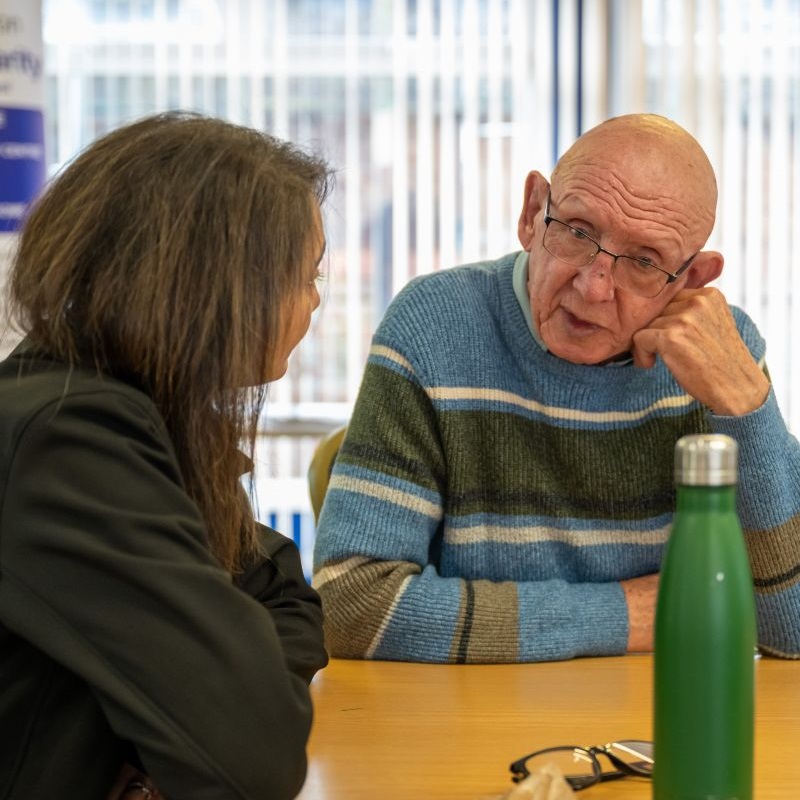 An older man with glasses and a striped sweater listens attentively to a woman at a table. The setting appears to be a community event with informational posters_image
