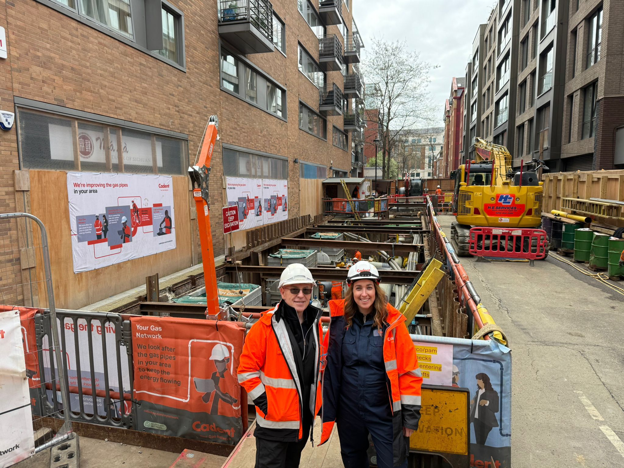 Two smiling construction workers in orange safety jackets and hard hats stand at an urban excavation site, with machinery and barriers behind them.