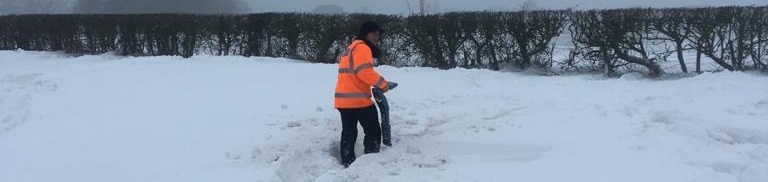 A person in an orange high-visibility jacket walks through deep snow in a foggy field, near a hedge. The scene is cold and wintry.