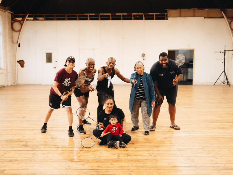 A diverse group of six adults and a child hold badminton rackets in a spacious hall, smiling and exuding a joyful and inclusive atmosphere.