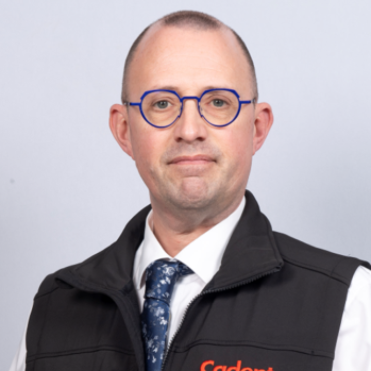 Portrait of David Garner wearing blue round glasses, a white shirt, patterned blue tie, and a black branded vest against a light gray background