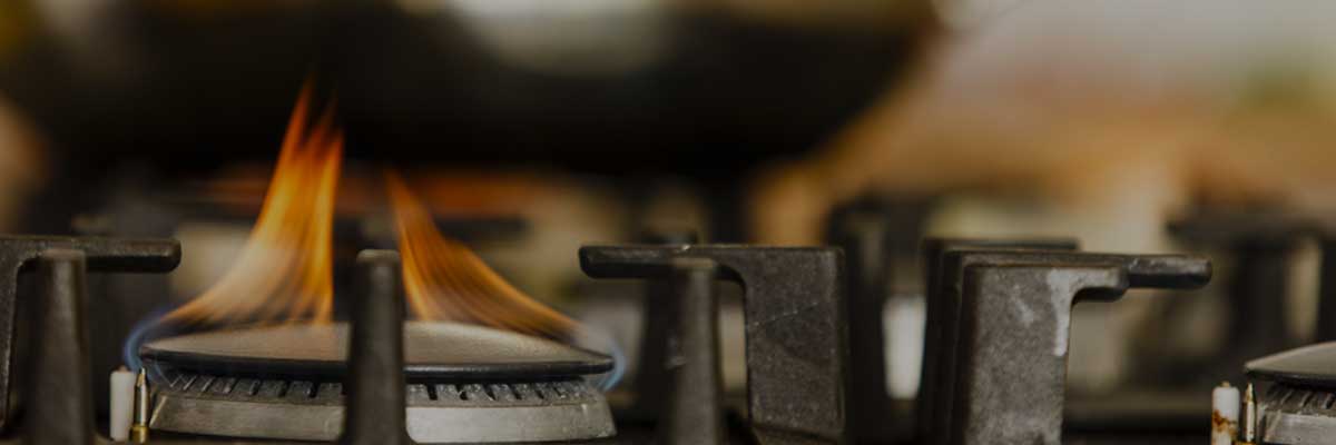 A close-up of a lit gas stove burner with visible blue and orange flames. The blurred background suggests a cozy kitchen atmosphere.