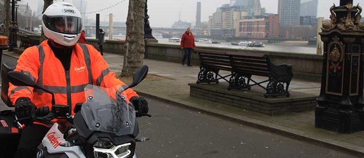 A motorcyclist in a bright orange jacket and helmet rides along a riverside path. Overcast day with trees, benches, and modern skyscrapers in the background.