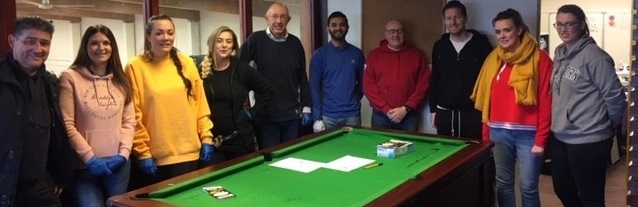 A diverse group of 10 people stand smiling around a green pool table in a dimly lit room. They wear casual clothing, creating a friendly, relaxed atmosphere.