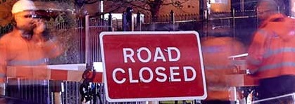 Workers in orange vests stand near a 'Road Closed' sign at night. Barriers and blurred motion convey a bustling, active work scene.