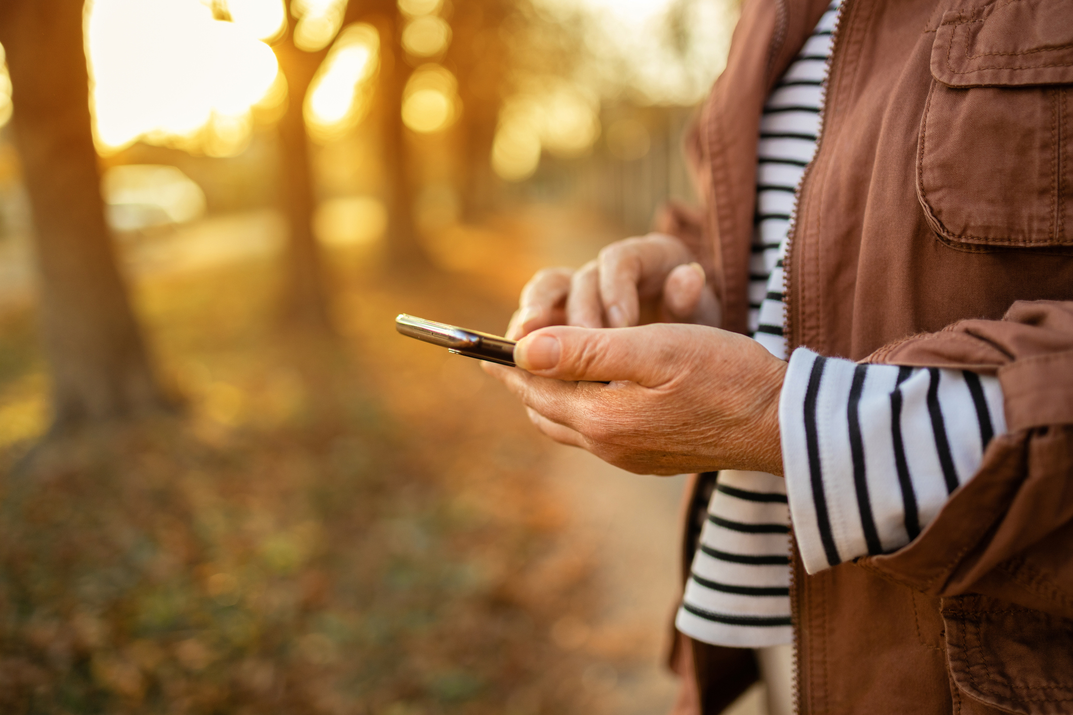 Person holding and using a smartphone while walking along a sunlit path lined with trees