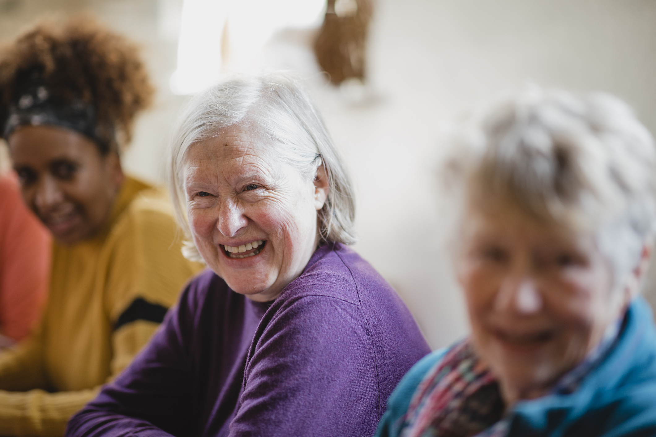 Three women are seated and smiling in a warmly lit room. The focus is on a woman in a purple sweater, conveying joy and camaraderie.