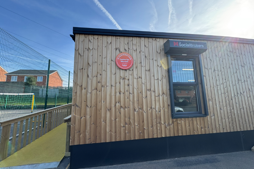 Wooden building with a 'Social Brokers' sign above a window. A red circular plaque is on the wall. Sunny sky with contrails and a fenced area.