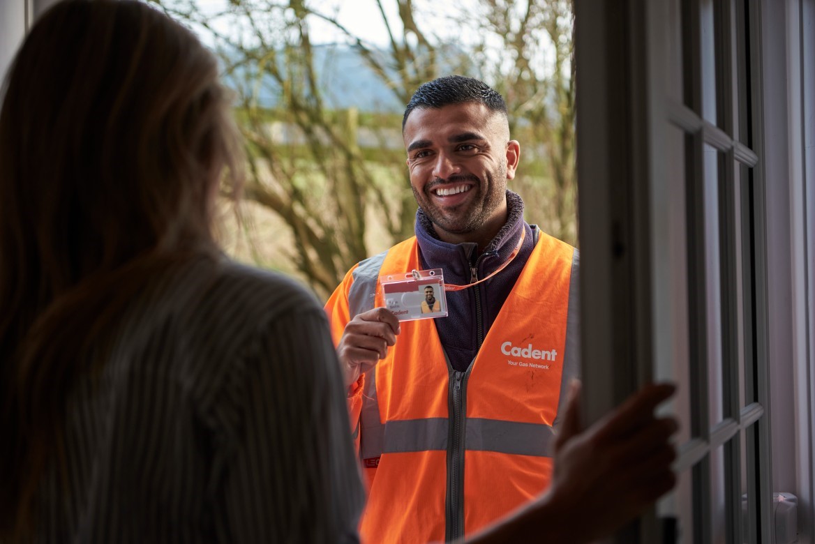 A man in an orange 'Cadent' vest holds an ID card, smiling warmly at a woman at her door. The scene conveys a friendly, professional visit.