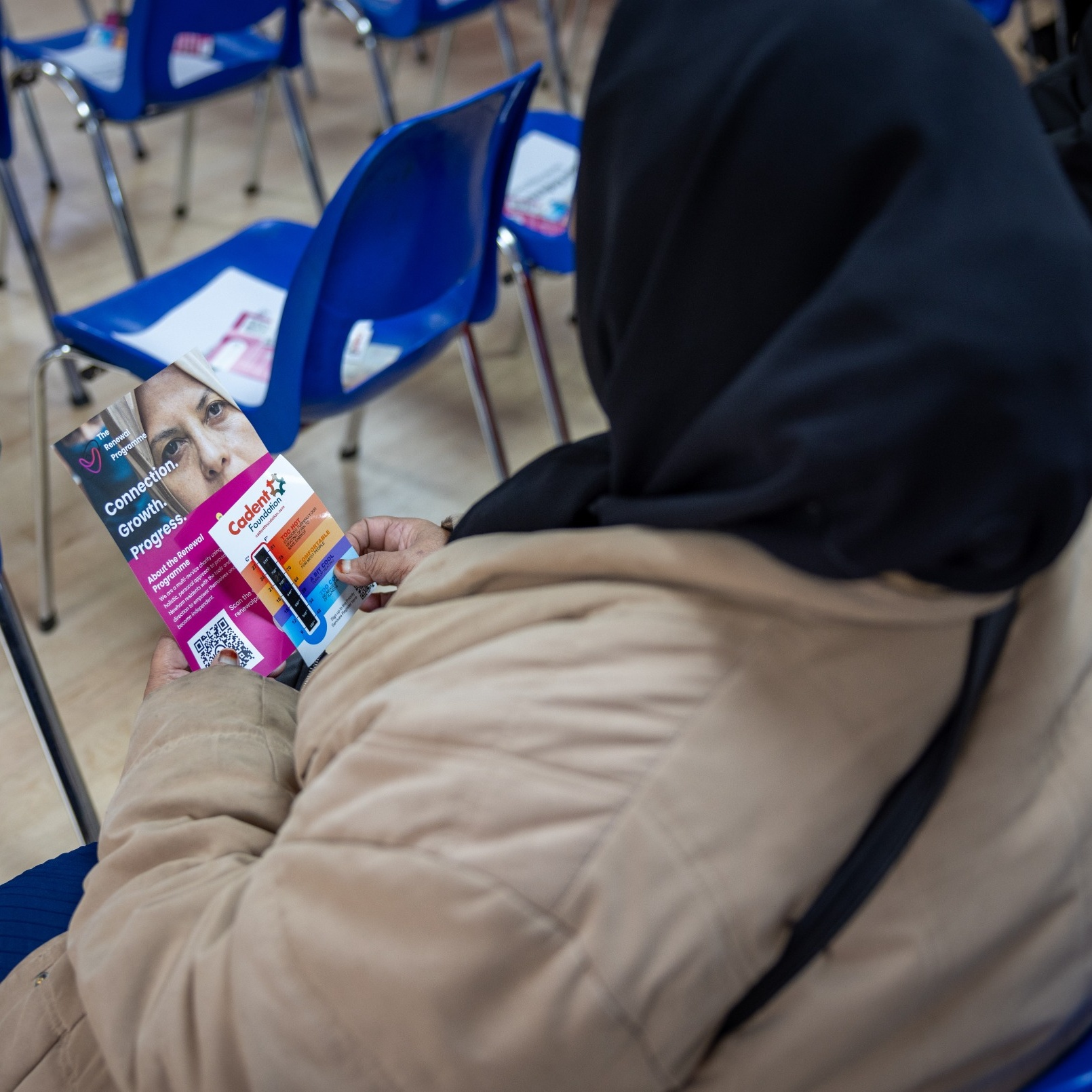 A person in a light brown coat and black headscarf holds a colorful pamphlet with text and a QR code. Blue chairs are arranged in rows in the background.