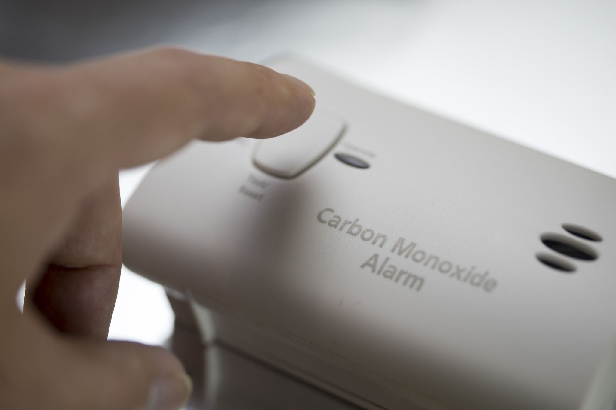 A hand is pressing the test button on a carbon monoxide alarm. The device is light gray with text indicating Carbon Monoxide Alarm, and has vents.