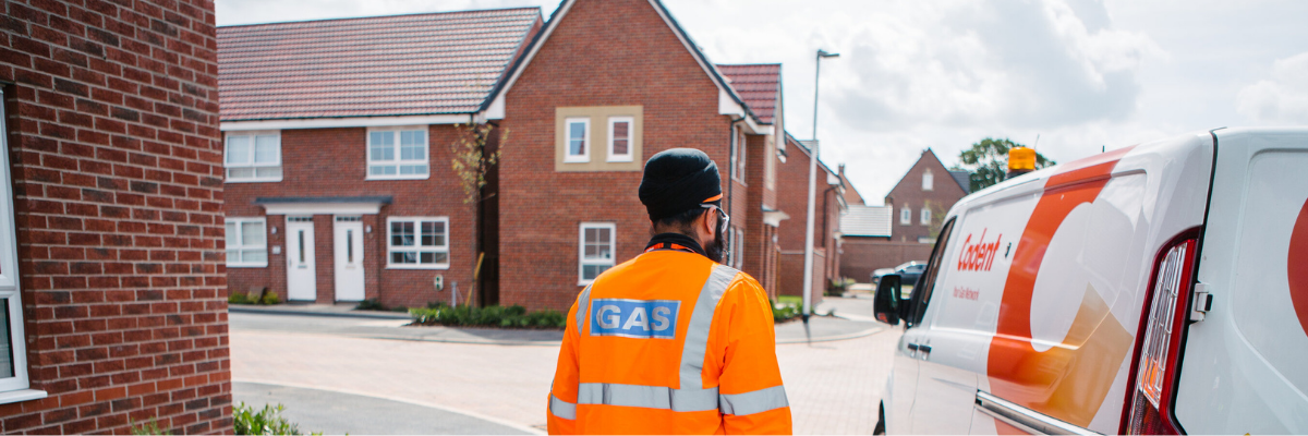 A gas engineer wearing a high-visibility orange jacket walks toward a residential street, with brick houses in the background and a service van parked nearby.