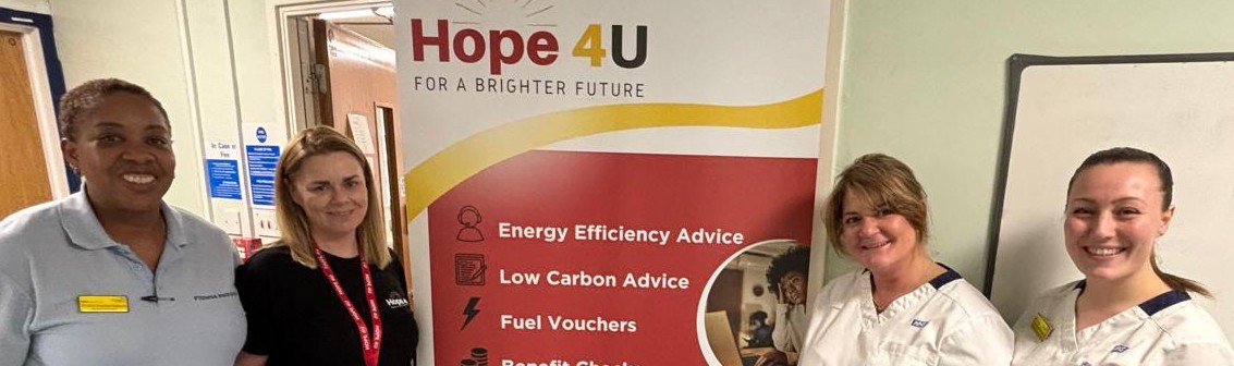 Four women stand beside a Hope 4U banner offering energy efficiency, low carbon advice, fuel vouchers, and benefit checks, in an indoor setting.