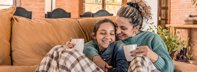A mother and daughter sit closely on a cozy sofa, sharing smiles and holding mugs. They're wrapped in a checkered blanket, conveying warmth and affection.