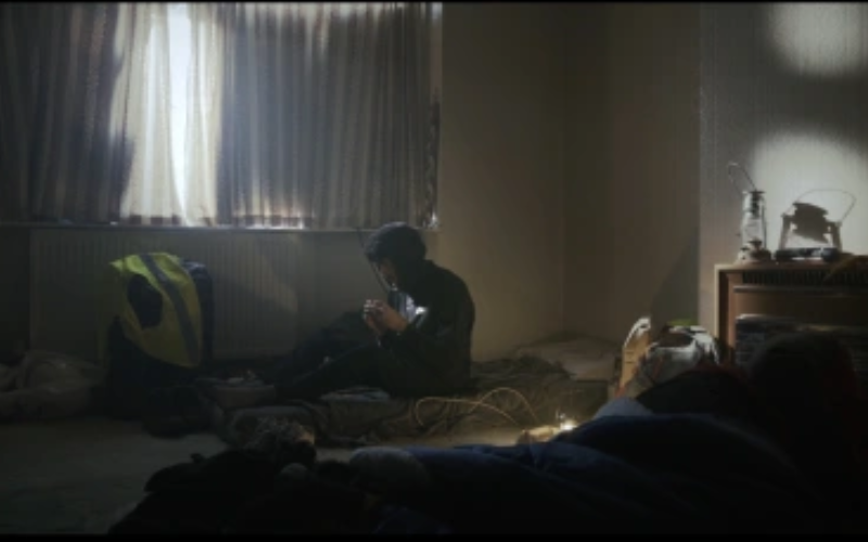 A dimly lit room with closed curtains allowing a small amount of daylight to filter through. A person wearing a hooded jacket sits on a thin mattress on the floor, looking down at something in their hands