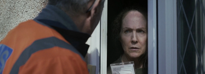 A woman cautiously looks out through a partially opened front door at a worker wearing an orange jacket, who is showing her an ID badge