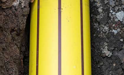 Close-up of a bright yellow pipe with brown stripes, partially buried in dark, rocky soil. The scene conveys construction or utility work.