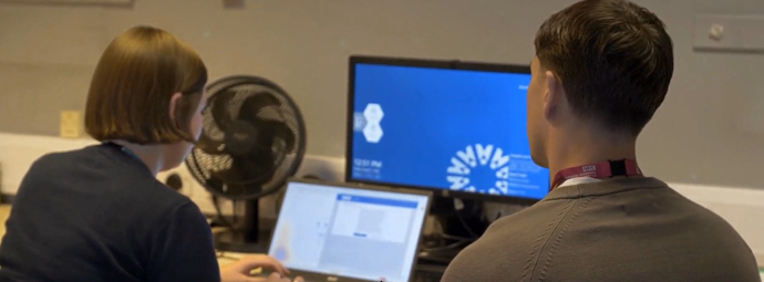 Two NHS staff collaborate at a desk, viewing a laptop and blue monitor in a hospital office; Manchester University NHS Foundation Trust logo visible.