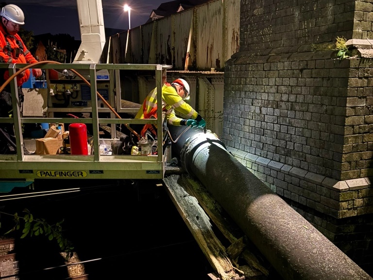 At night, two hard-hat workers in high-visibility gear guide a large pipe from a raised platform beside a stone wall during industrial repairs.