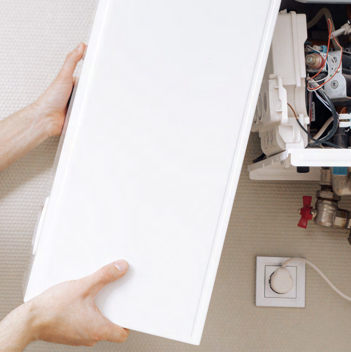 Hands holding a white cover in front of an open boiler with exposed components and wires. A power outlet and plug are visible on the wall nearby.