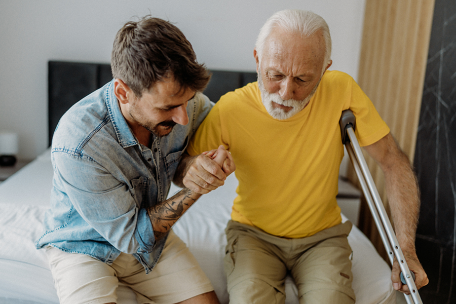 A young man supports an older male adult using a crutch while helping them move from a bed.