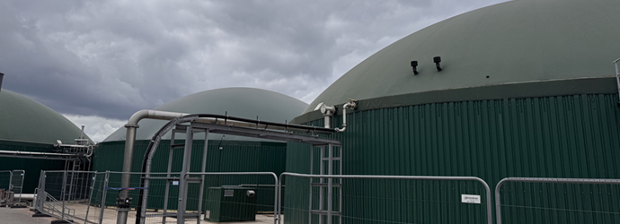 Large green domed industrial tanks behind metal security fencing, with pipes and gates under a dark, cloudy sky, creating a somber atmosphere.