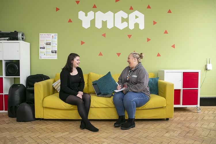 Two women sit on a yellow sofa in a room with a green wall featuring the YMCA logo. They are smiling and talking, creating a friendly and welcoming atmosphere.