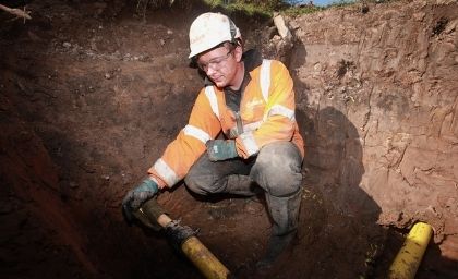 A worker in an orange safety jacket and helmet is crouching in a trench, inspecting a yellow pipe. The scene conveys focus and diligence.