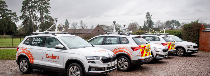 A row of four white utility vehicles parked on a gravel driveway, each marked with orange and red safety graphics and the word “Cadent.” 