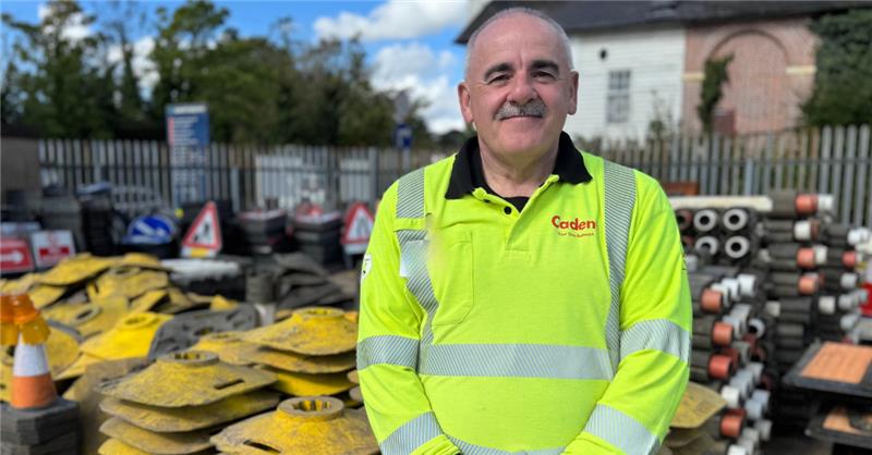 A man in a high-visibility yellow jacket stands confidently in a construction yard with stacked pipes and signs around. The scene is bright and industrial_image