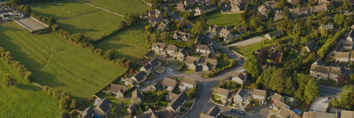 Aerial view of a quaint village with neatly arranged houses interspersed with greenery. Sunlit fields surround the village, adding a serene and peaceful vibe.