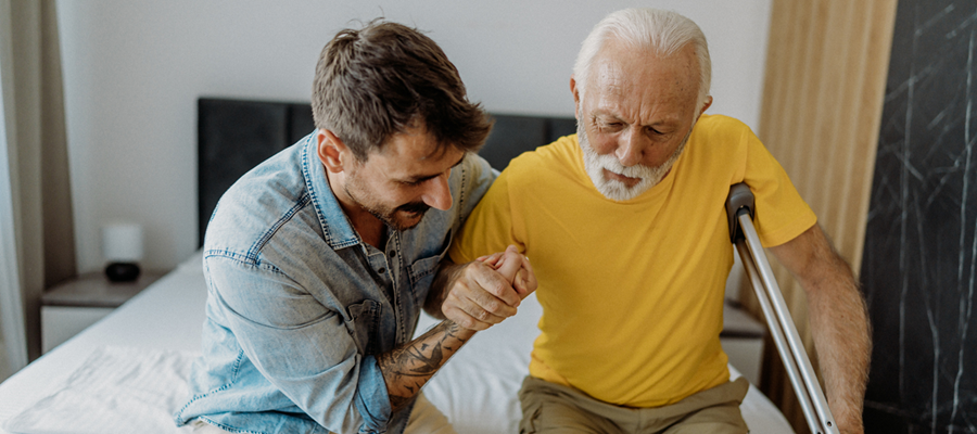 A person helps an older adult with a walking aid sit up on the edge of a bed, providing physical support in a home setting.