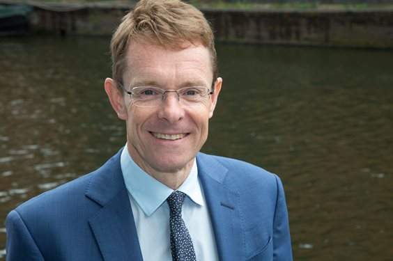 A man in a blue suit and glasses smiles warmly against a backdrop of calm water. The setting conveys a professional yet relaxed atmosphere.