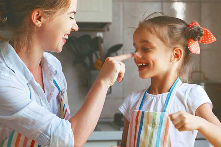 A woman and girl in striped aprons, smiling in a kitchen. The woman playfully touches the girl's nose with flour, creating a joyful, playful atmosphere.