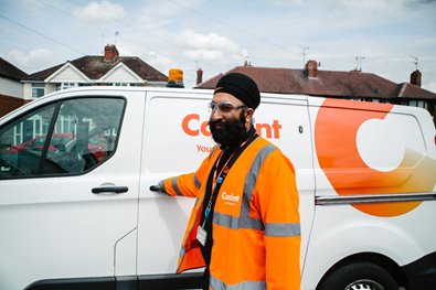 A man in an orange safety jacket stands beside a white utility van with a logo, on a suburban street. He appears focused and professional.