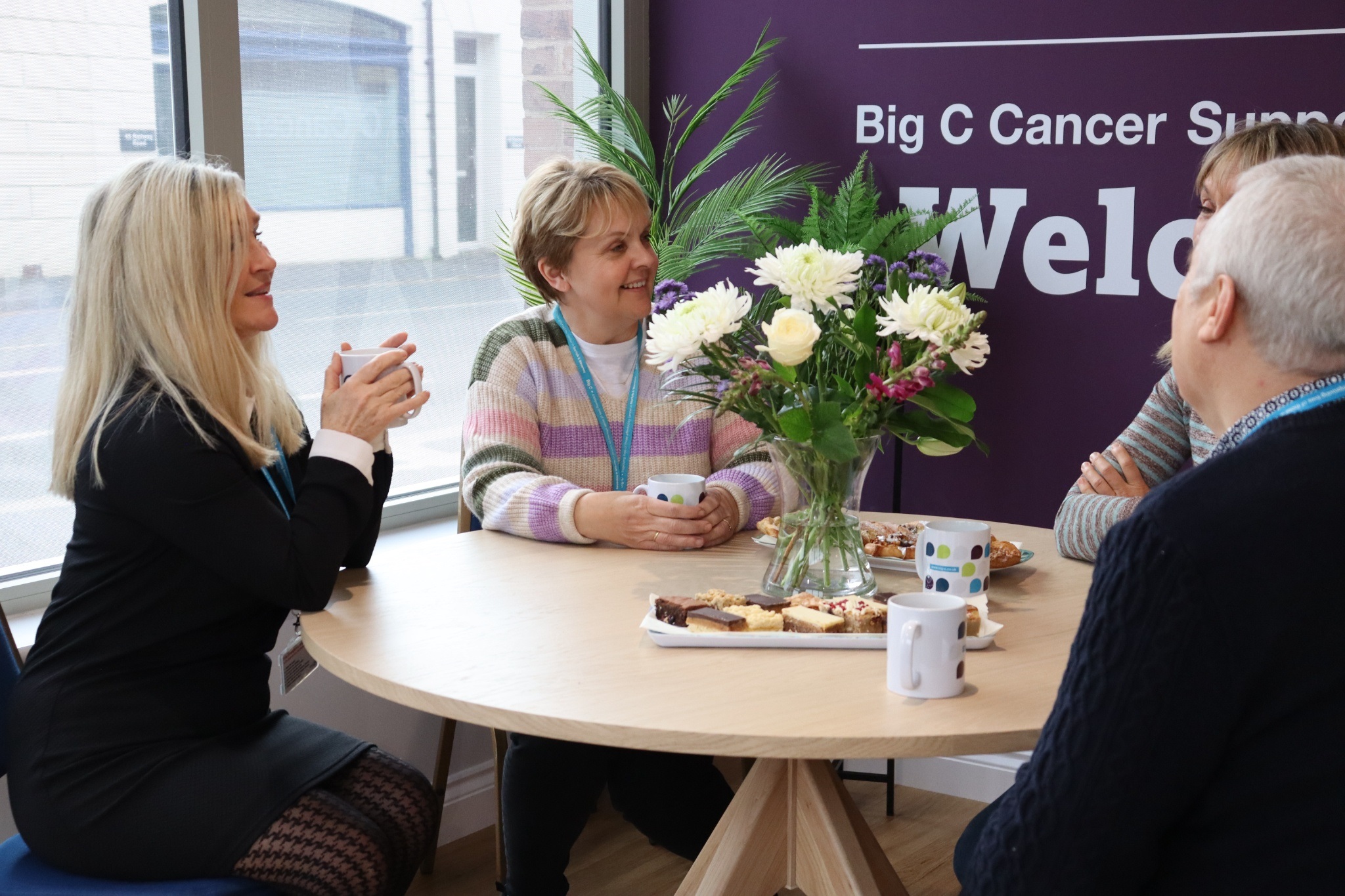Four people sit around a table with mugs and pastries, smiling warmly. A vase of flowers is at the center. A purple wall reads 'Big C Cancer Support Welcome.'