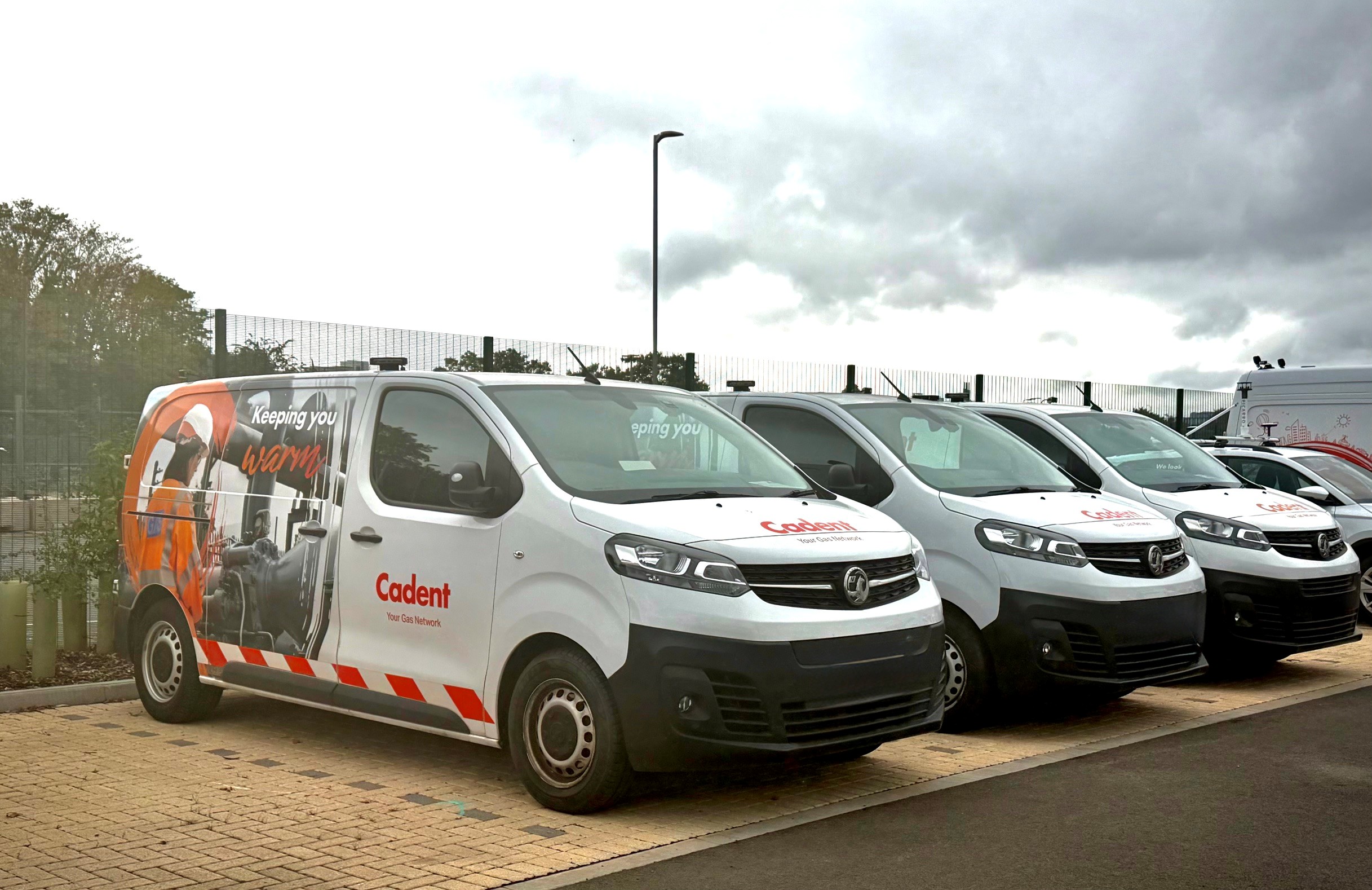 Four white Codent service vans, with orange graphics, are parked in a row outside under a dark, cloudy sky.