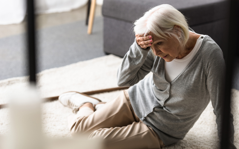 Elderly woman sitting on the floor looking unwell and holding her head.