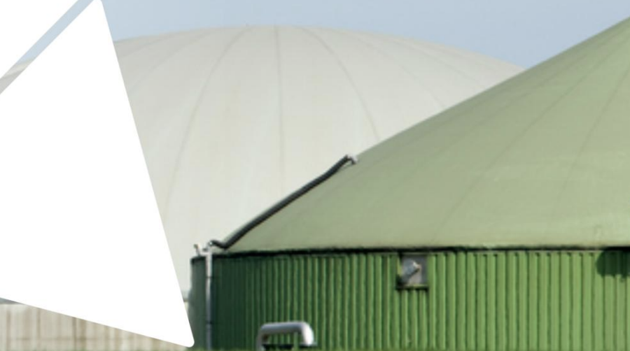 A green cylindrical industrial tank with a sloped roof, pipes along the top edge, and a large white domed structure visible in the background against a clear sky.