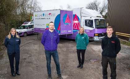 Four people stand in front of a large truck and a van with colorful branding on a gravel path, smiling, conveying a team spirit and positivity.
