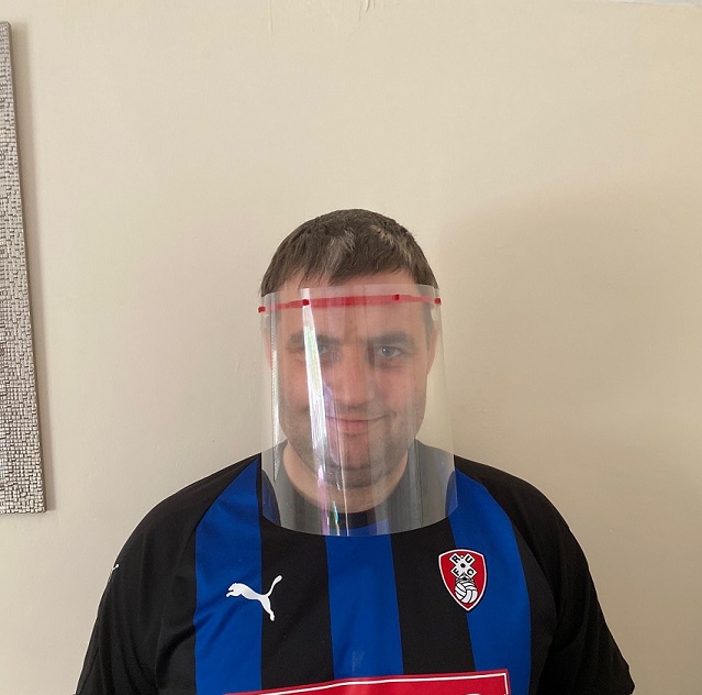 Man wearing a face shield, smiling slightly, dressed in a black and blue striped sports jersey with a logo. Neutral beige background.