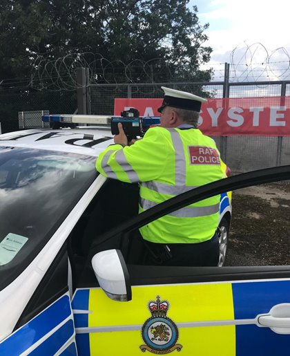 RAF police officer in a high-visibility jacket uses a speed radar from a patrol car. A fence with barbed wire is in the background under a cloudy sky.